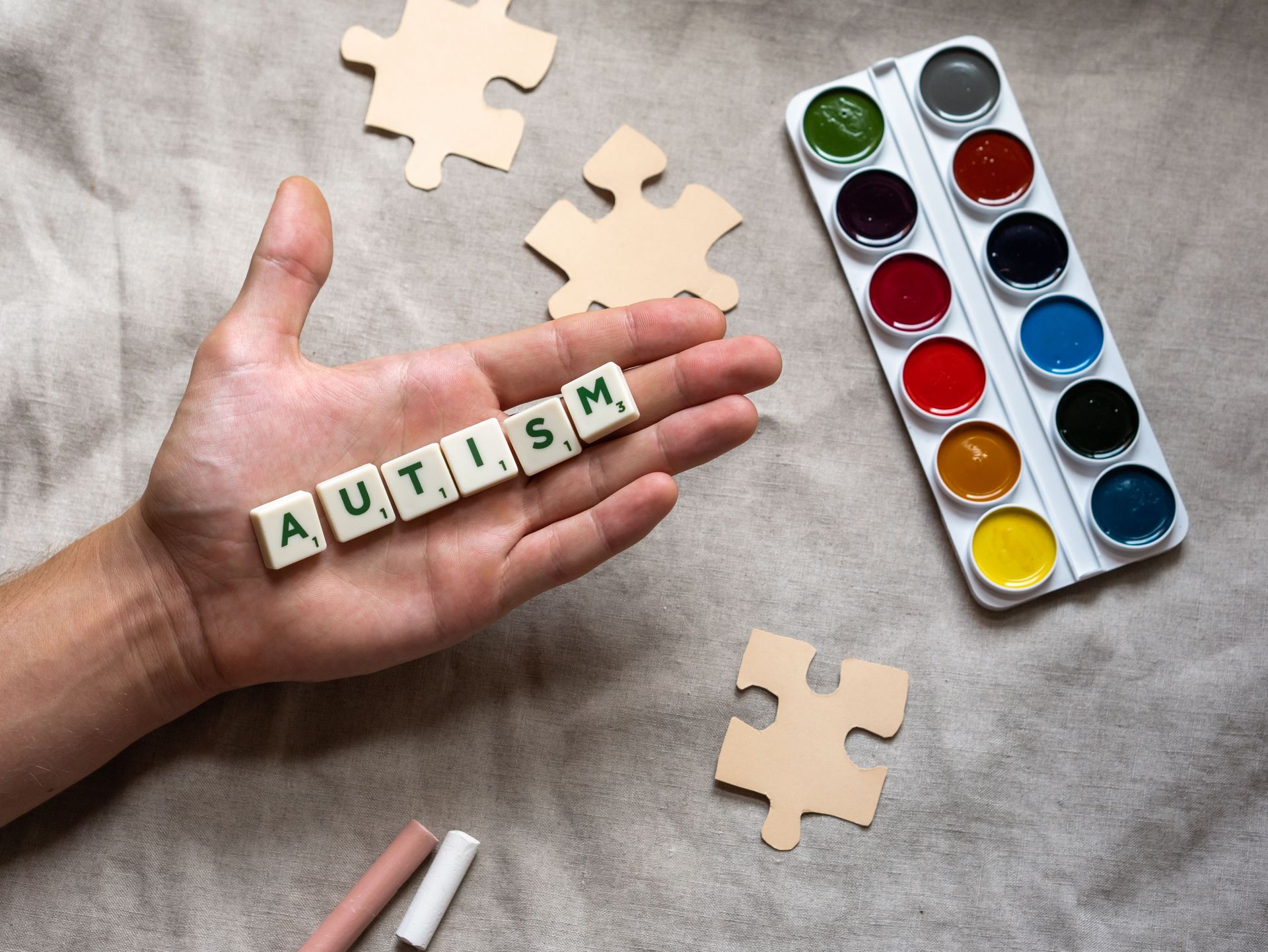 Hand holding autism scrabble tiles surrounded by puzzle pieces and watercolor paints, promoting autism awareness.