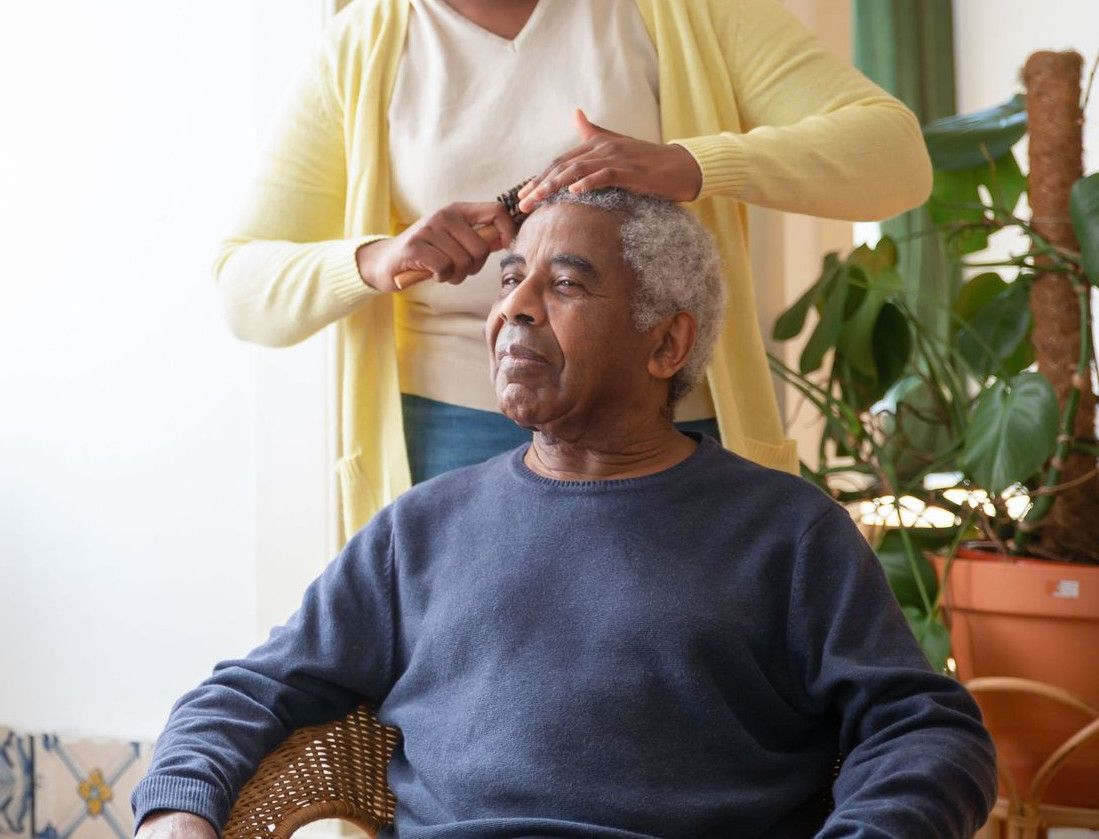 A caregiver combing an elderly man's hair near a sunlit window indoors.