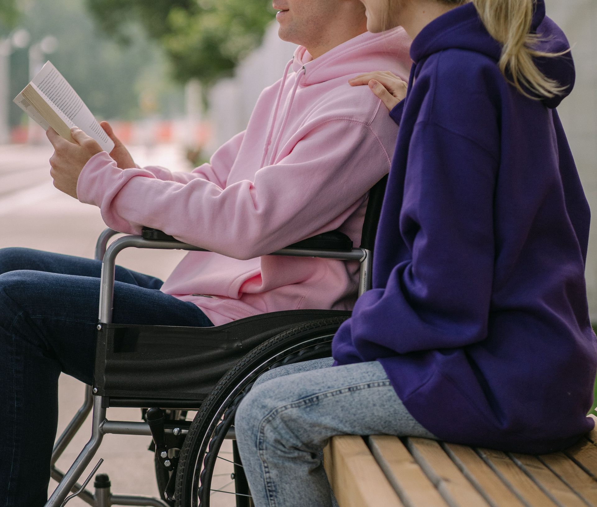A young man in a wheelchair reading a book with a friend outdoors on a sunny day.