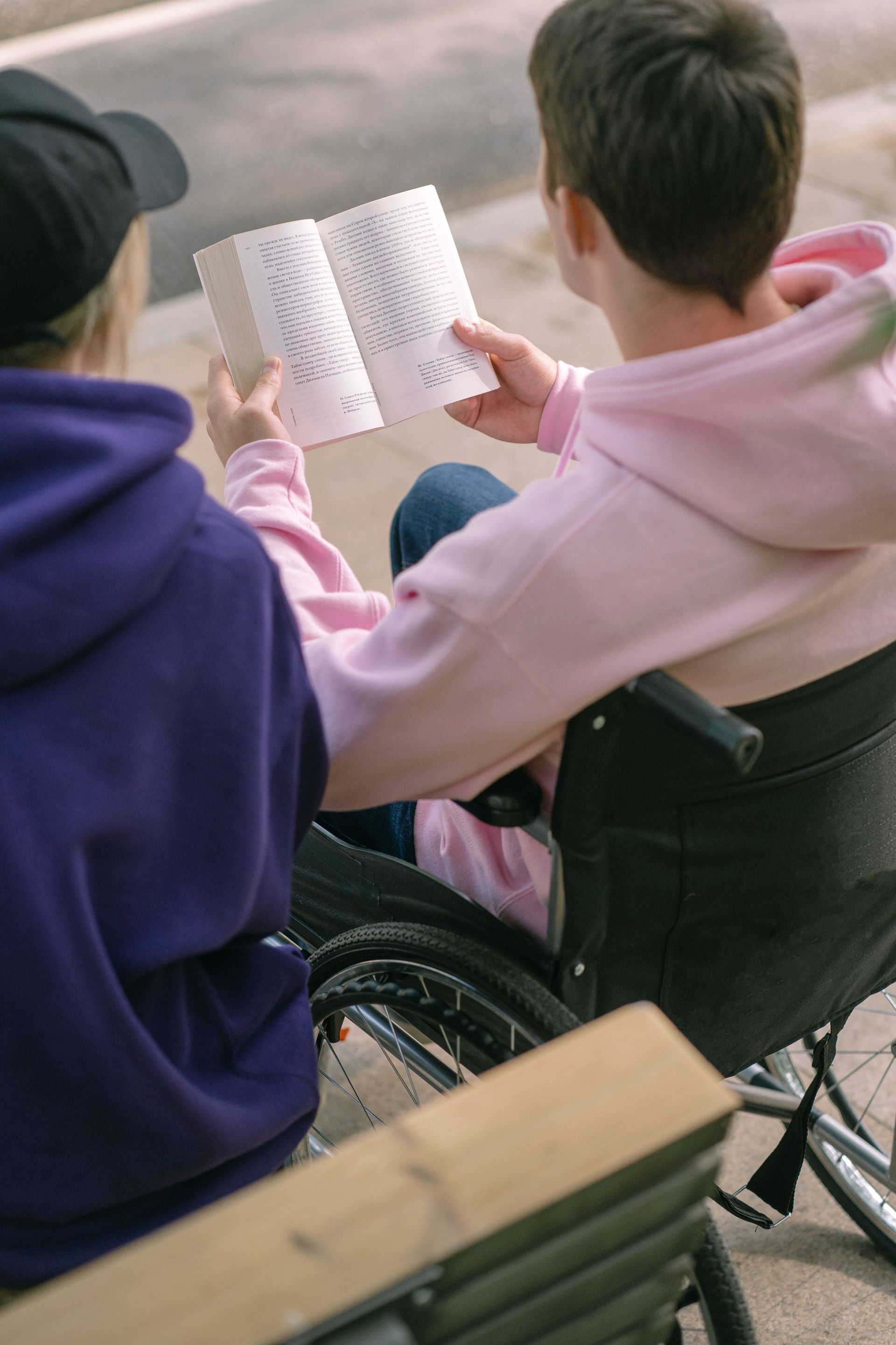 A young man in a wheelchair enjoys reading a book outdoors with a friend beside him.